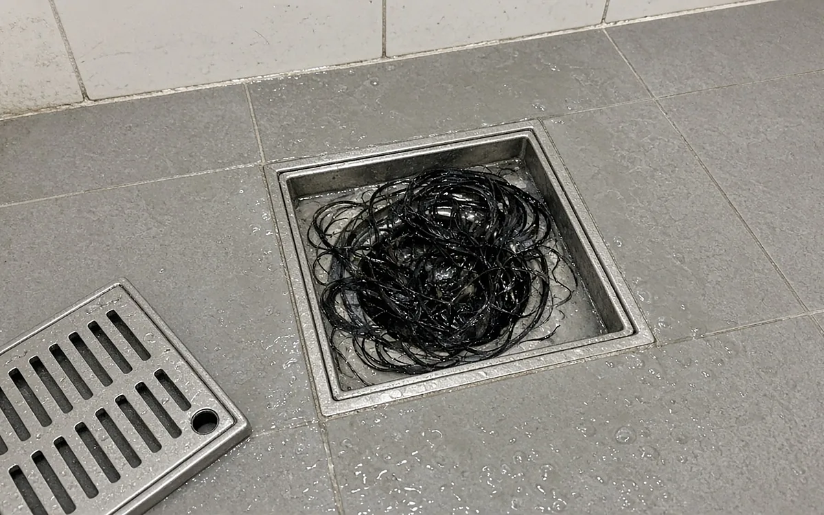 Thoát sàn inox vuông trong nhà tắm lát gạch xám, mặt lưới đã tháo đặt bên cạnh, bên trong là một búi tóc đen dài quấn chặt bít gần kín miệng thoát, nước đọng lấm tấm trên sàn