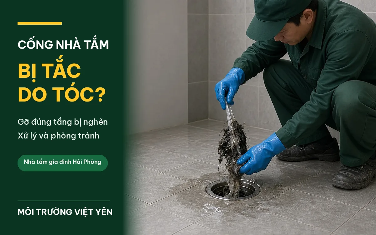 Kỹ thuật viên mặc đồng phục xanh đậm, găng tay xanh dương ngồi xổm kéo búi tóc lẫn cặn bẩn ra khỏi thoát sàn inox trong phòng tắm lát gạch xám, ảnh bìa cống nhà tắm bị tắc do tóc