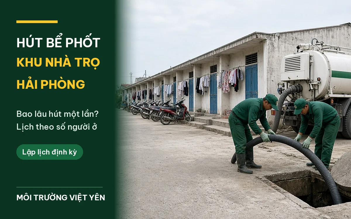 Ban ngày trong lối đi dãy nhà trọ một tầng cửa xanh, xe máy dựng dọc hiên và quần áo phơi, hai kỹ thuật viên đồng phục xanh đậm luồn ống hút đen vào bể phốt bê tông đang mở, xe hút trắng sọc xanh đỗ bên phải, ảnh bìa hút bể phốt nhà trọ Hải Phòng