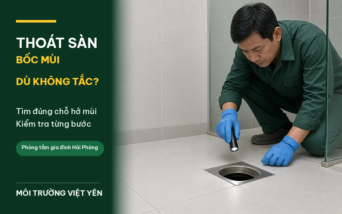 Kỹ thuật viên mặc đồng phục xanh đậm, găng tay xanh dương quỳ trên sàn gạch sáng màu, rọi đèn pin vào thoát sàn inox vuông đã tháo mặt lưới để tìm nguồn mùi trong phòng tắm, ảnh bìa thoát sàn phòng tắm bốc mùi