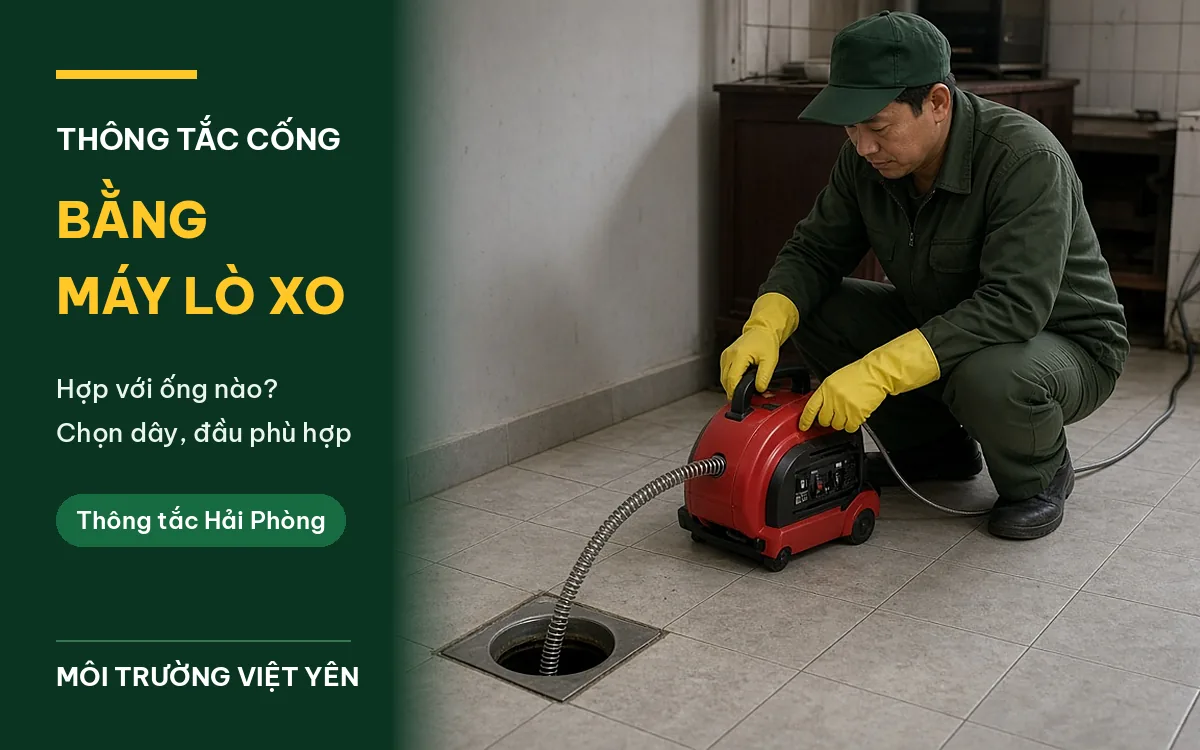Kỹ thuật viên mặc đồng phục xanh lá, đội mũ lưỡi trai, đeo găng tay cao su vàng ngồi trên sàn lát gạch, đưa dây cáp thép của máy thông cống lò xo chạy điện màu đỏ vào miệng thoát sàn vuông, ảnh bìa bài thông tắc cống bằng máy lò xo